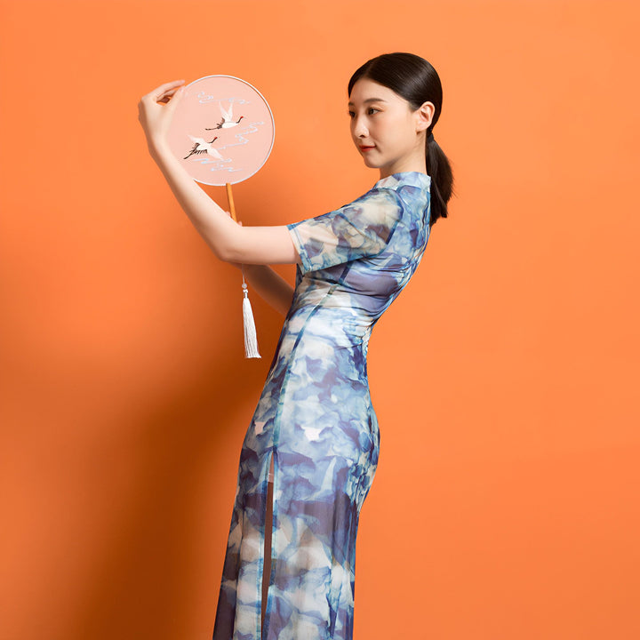 Femme en kimono bleu à motifs floraux tenant un éventail orné de grues. Élégance et tradition dans notre collection de kimonos. Découvrez nos créations uniques.