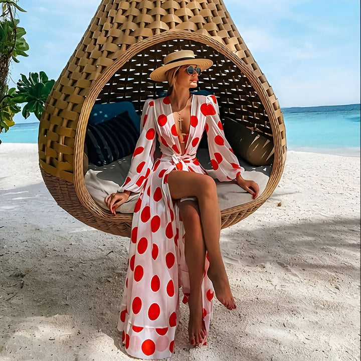 Femme élégante en kimono blanc à pois rouges, assise dans une balançoire en osier sur une plage de sable blanc. Mer turquoise en arrière-plan. Kimonos tendances pour femmes.