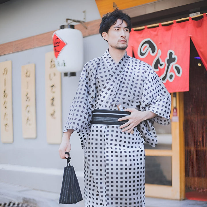 Homme portant un kimono traditionnel japonais à motifs, avec une ceinture obi noire. Il tient un petit sac, devant une boutique de style japonais. Kimonos authentiques à vendre.