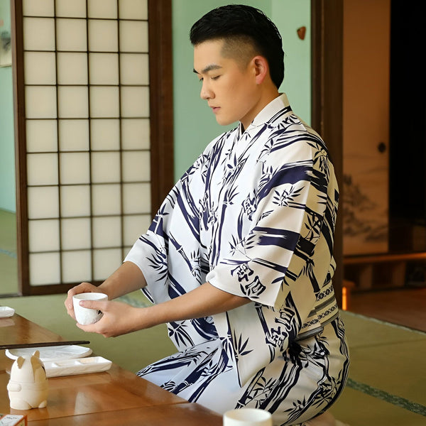 Homme asiatique portant un kimono blanc et bleu marine avec motif de bambou. Il est assis à une table en bois avec une tasse et des plats. Kimonos disponibles à l'achat.
