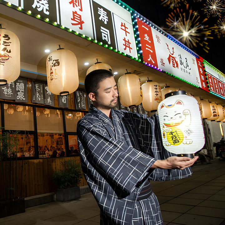Homme en kimono tenant une lanterne Maneki Neko devant un restaurant japonais illuminé. Vêtements traditionnels japonais en vente.