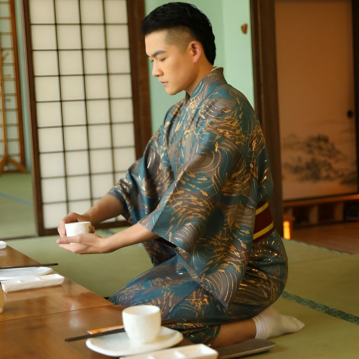 Homme en kimono bleu à motifs dorés, assis à genoux dans un intérieur japonais traditionnel avec un service à thé. Vente de kimonos pour hommes.